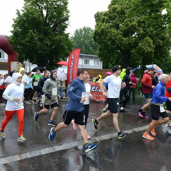 Läufer in verschiedenen farbigen Klamotten nehmen an einem Marathon im Regen teil. Ein Mann in einem blauen Jackett hält ein Papier. Bäume und ein roter Banner sind an der Seite.