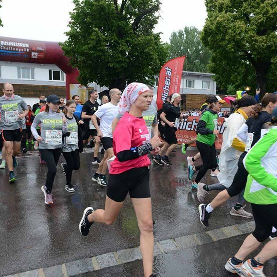 Eine Gruppe von Menschen nimmt an einem Marathon teil und läuft in einer Reihe auf einer nassen Straße. Einige tragen nummerierte Startnummern, und eine Frau in einem rosa Shirt führt an. Bäume und Gebäude säumen die Straße.