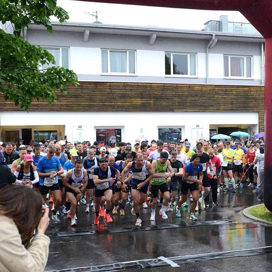 Eine Gruppe von Läufern nimmt an einem Marathon auf einer nassen Straße teil. Zuschauer beobachten von den Seitenlinien, und einige stehen unter Regenschirmen. Ein Gebäude mit mehreren Fenstern ist im Hintergrund.