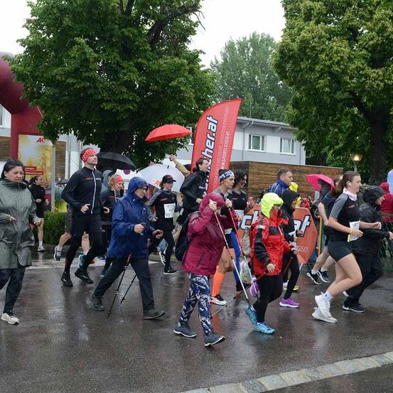 Marathonteilnehmer laufen auf einer nassen Straße. Viele tragen Regenschirme und Regenmäntel. Ein roter Banner mit weißen Texten ist im Hintergrund sichtbar.