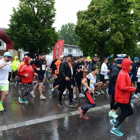Läufer in verschiedenen farbigen Ausrüstungen nehmen an einem Marathon auf einer nassen Straße teil, mit Bäumen und Gebäuden im Hintergrund.