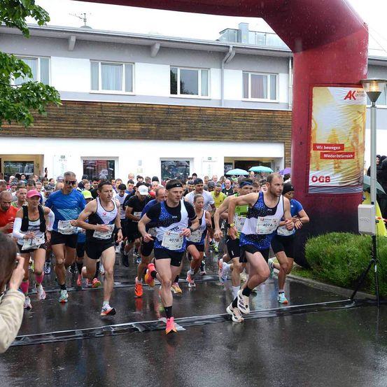 Eine Gruppe von Läufern nimmt an einem Marathon bei regnerischem Wetter teil. Zuschauer beobachten von den Seitenlinien. Im Hintergrund sind Gebäude und ein Schild sichtbar.