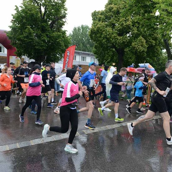 Eine Gruppe von Läufern nimmt an einem Marathon bei Regen teil. Sie tragen Turnschuhe und Shirts. Einige benutzen Regenschirme. Im Hintergrund sind Bäume und ein Gebäude zu sehen.