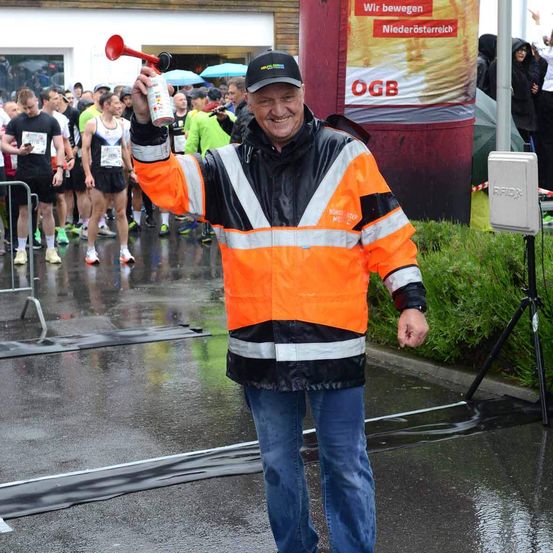Ein lächelnder Mann in einer reflektierenden orangefarbenen Weste und Jeans bläst eine Hupe, während er bei einem Marathon im Regen steht.