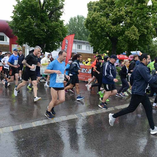 Eine Gruppe von Läufern nimmt an einem Marathon auf einer nassen Straße teil, einige tragen Regenmäntel. Ein Banner mit einer roten Flagge ist im Hintergrund sichtbar. Bäume und ein Gebäude sind im Hintergrund zu sehen.