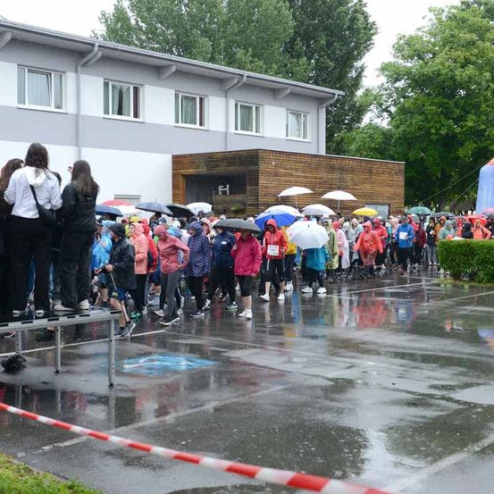 Teilnehmer stehen in einer nassen Parkbucht Schlange, halten Regenschirme, tragen Regenmäntel und Turnschuhe. Das Gebäude im Hintergrund hat mehrere Fenster und einen hölzernen Anbau.