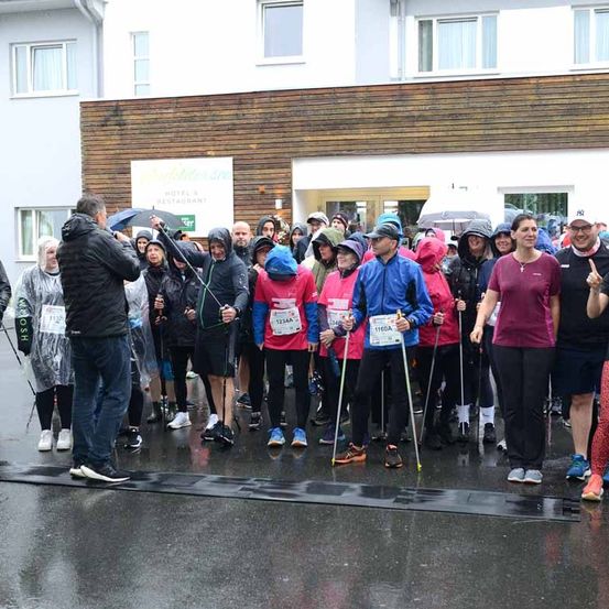 Marathon-Teilnehmer bei Regen stehen vor einem Gebäude, einige mit Regenschirmen, in Regenjacken und Turnschuhen, halten Skistöcke.