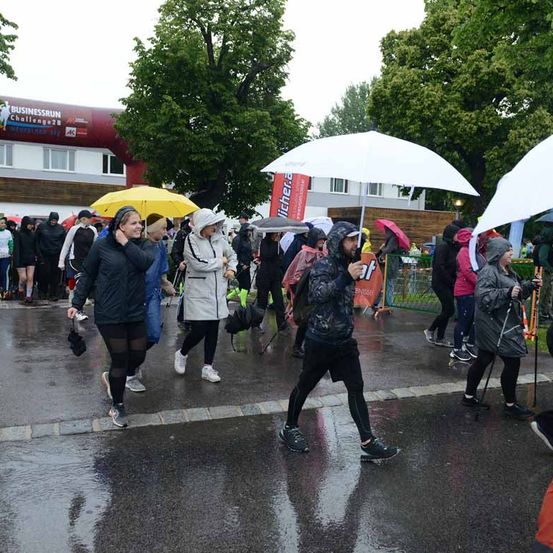 Menschen gehen an einem regnerischen Tag auf einer nassen Straße. Einige tragen Regenschirme und andere Regenmäntel. Bäume und ein Gebäude mit einem Schild sind im Hintergrund.