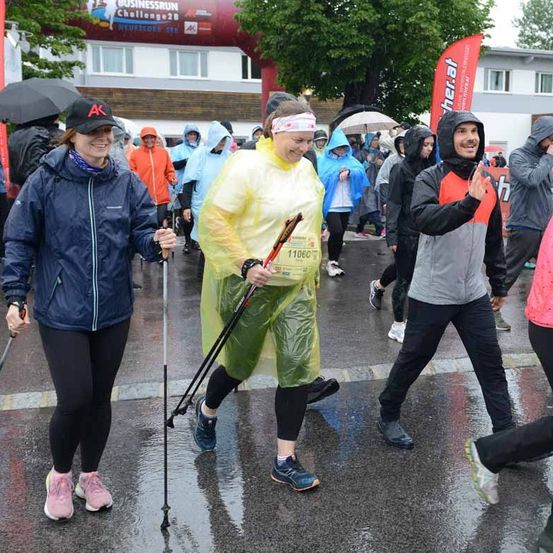 Läufer nehmen an einem Marathon bei regnerischem Wetter teil, tragen Regenmäntel und halten Trekkingstöcke. Sie laufen auf einer nassen Straße in der Nähe eines Gebäudes mit einem Banner.