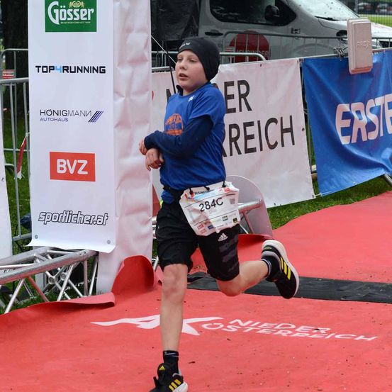 Ein junger Junge läuft einen Marathon, trägt ein blaues Shirt und schwarze Shorts. Er hat die Nummer 284C auf seinem Shirt. Hinter ihm sind Banner mit dem Text "ER REICH" und "ERSTE". Ein Van ist hinter dem Zaun geparkt.