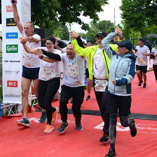 Eine Gruppe von Läufern mit Nummern und Turnschuhen feiert an der Ziellinie eines Marathons. Sie heben die Hände und lächeln. Der Boden ist mit rotem Teppich bedeckt.