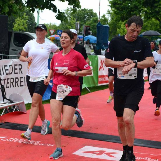 Läufer bei einem Marathonrennen auf nassem Boden, eine Frau in einem roten Shirt läuft vorne, und ein Mann in Schwarz überprüft seine Uhr.