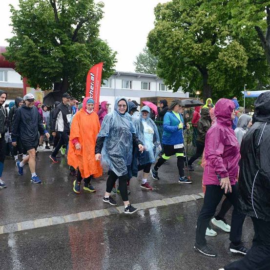 Marathonteilnehmer, einige mit Regenschirmen, gehen auf einer nassen Straße. Der Himmel ist bedeckt.