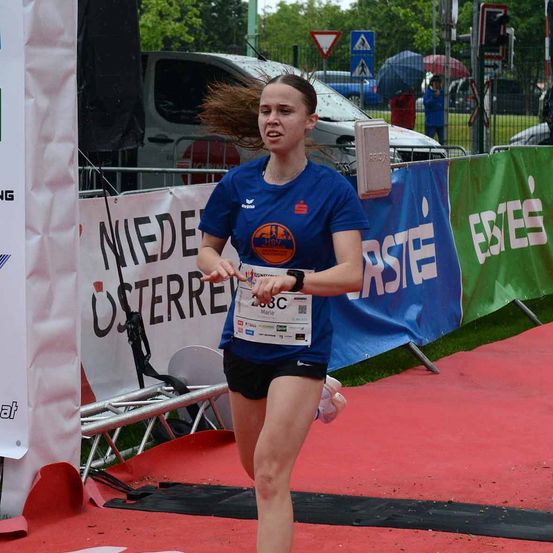 Eine Frau läuft auf einer roten Laufbahn mit einem Nummernschild, trägt ein blaues T-Shirt und schwarze Shorts. Hinter ihr sind Banner und ein Zaun zu sehen, zusammen mit Menschen unter Regenschirmen.