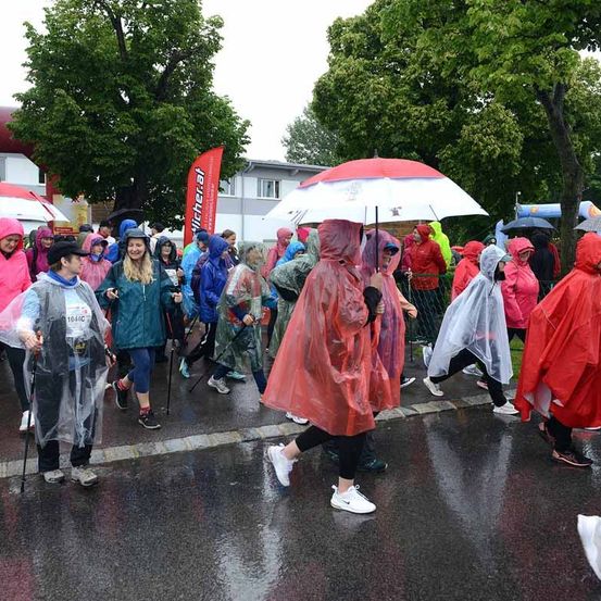 Marathonteilnehmer in Regenmänteln gehen unter Regenschirmen auf einer nassen Straße. Bäume und Gebäude säumen die Straße, mit einem Zaun und Bannern im Hintergrund.