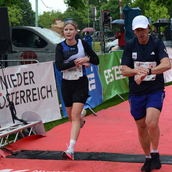 Eine Frau und ein Mann nehmen an einem Marathon teil. Die Frau mit der Startnummer 4088 läuft auf einem roten Teppich. Der Mann mit der weißen Kappe hält eine Uhr. Sie stehen neben einem Banner mit dem Text 'Nieder Osterreich'. Im Hintergrund steht ein Lieferwagen und ein Lautsprecher.