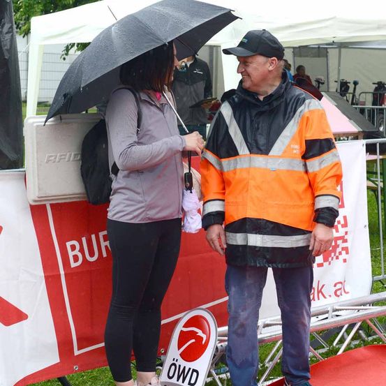 Eine Frau mit einem Regenschirm steht neben einem Mann in einer reflektierenden orangefarbenen Weste. Sie lächeln beide und schauen sich an. Hinter ihnen befindet sich ein weißes Banner.