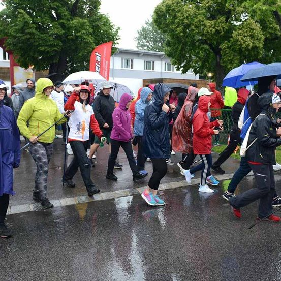 Teilnehmer eines Marathons, die Regenjacken tragen und Regenschirme halten, laufen auf einer nassen Straße. Ein Banner ist links zu sehen, und Bäume und Gebäude sind im Hintergrund.