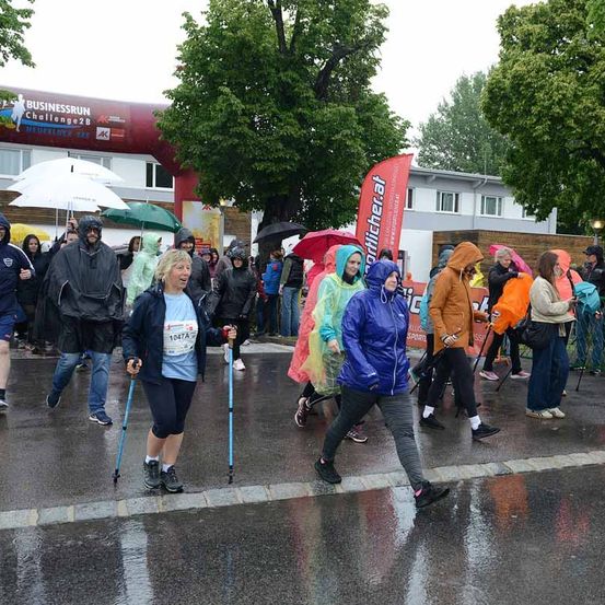 Eine Gruppe von Menschen läuft auf einer nassen Straße bei einem Marathon. Sie tragen Regenmäntel und halten Regenschirme. Ein Banner wird von einem der Teilnehmer gehalten. Ein Baum und ein Gebäude befinden sich am Straßenrand.