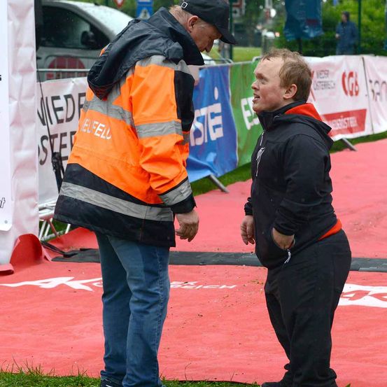 Zwei Männer stehen sich auf einer roten Laufbahn gegenüber, einer in einer reflektierenden orangefarbenen Jacke und der andere in einer schwarzen Kapuzenjacke. Hinter ihnen werben Banner mit dem Wort 'STEL' und anderem Text.