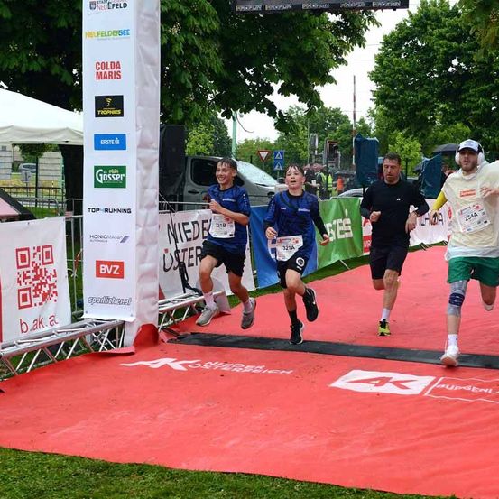 Eine Gruppe von Läufern überquert die Ziellinie eines Marathons auf einem roten Teppich, mit Werbebannern an den Seiten. Ein Läufer hält ein Papier in der Hand, und andere tragen Ohrhörer.