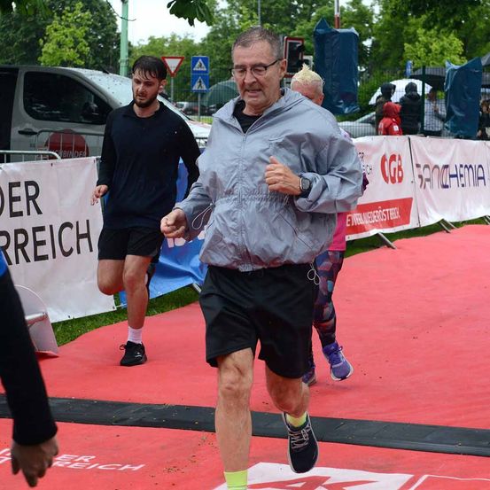 Ein älterer Mann läuft auf einer roten Laufbahn, trägt eine Brille und eine Jacke, mit einem jüngeren Mann, der hinter ihm läuft. Sie nehmen wahrscheinlich an einem Marathon teil.