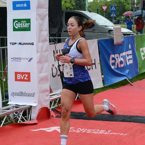 Eine Frau läuft auf einer roten Laufbahn bei einem Rennen. Hinter ihr sind Banner mit Logos und Texten. Autos und Menschen sind im Hintergrund.