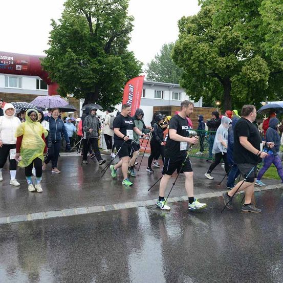 Teilnehmer eines Marathons laufen bei Regen, halten Regenschirme und tragen Regenmäntel. Ein roter Banner ist im Hintergrund zu sehen.