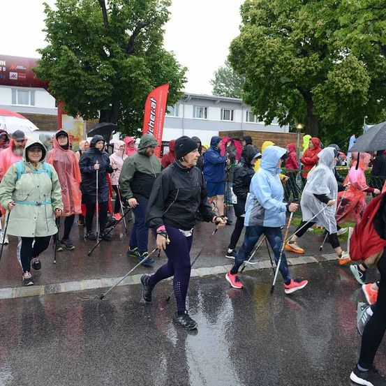 Eine Gruppe von Menschen geht bei Regen mit Regenschirmen und Regenmänteln. Sie halten Stöcke und einige haben Hüte auf. Bäume und ein Gebäude sind im Hintergrund.