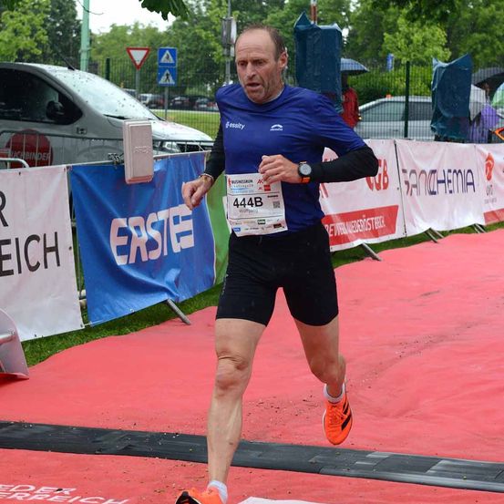 Ein Mann läuft in einem Marathon. Er trägt ein blaues T-Shirt mit der Nummer 444B und schwarze Shorts. Er hat eine orangefarbene Armbanduhr. Hinter ihm stehen Banner, und einige Leute stehen unter Regenschirmen.