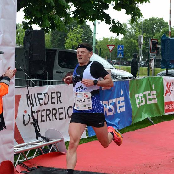 Ein Mann läuft einen Marathon und trägt die Nummer 407B auf seinem Trikot. Er trägt einen Stirnband und eine Uhr. Eine orange Person ist auf seiner linken Seite, und ein weißer Banner mit den Worten 'Nieder Osterreich' ist auf seiner rechten Seite.