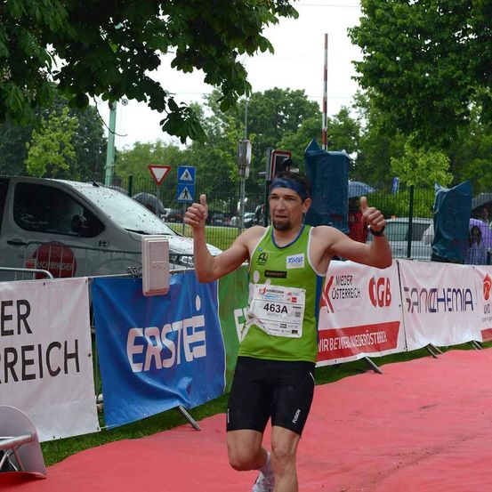 Ein Mann in einem grünen Trikot mit der Nummer 463A läuft auf einer roten Bahn und jubelt mit beiden Händen. Dahinter sind Banner mit Erste und anderen Texten. Ein Van ist auf der Seite geparkt.