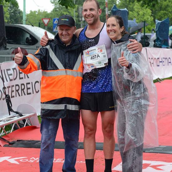 Ein Läufer in einem blau-weißen Singlet mit der Nummer 407A steht zwischen zwei Personen auf einer roten Laufbahn, lächelt und gibt ein Daumen hoch.