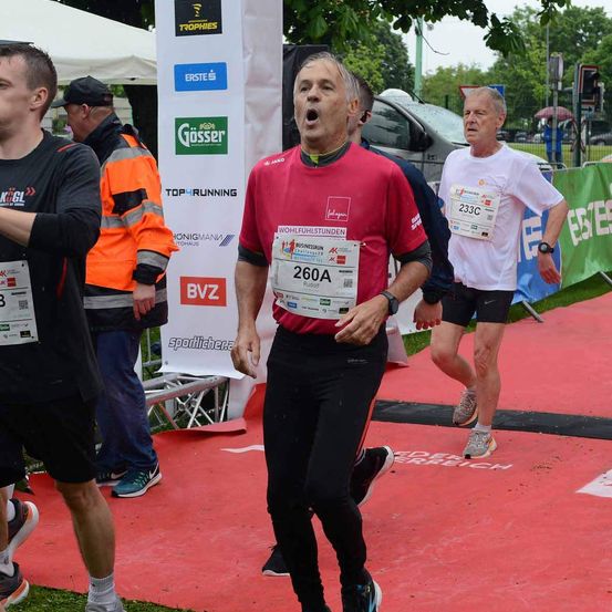 Eine Gruppe von Menschen läuft bei einem Marathon. Ein Mann in einem roten Shirt trägt die Nummer 260A. Hinter ihm ist ein Mann in einem weißen Shirt mit der Nummer 233C. Sie laufen auf einer roten Laufbahn.