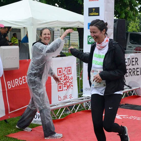 Zwei Frauen laufen und lachen, eine in einer Regenjacke und die andere in einer schwarzen Jacke. Sie befinden sich an der Ziellinie eines Marathons.
