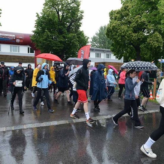 Eine Gruppe von Menschen geht bei Regen, trägt Regenmäntel und hält Regenschirme. Sie nehmen wahrscheinlich an einem Marathon teil, mit einem Gebäude im Hintergrund und einem roten Bogenschild.