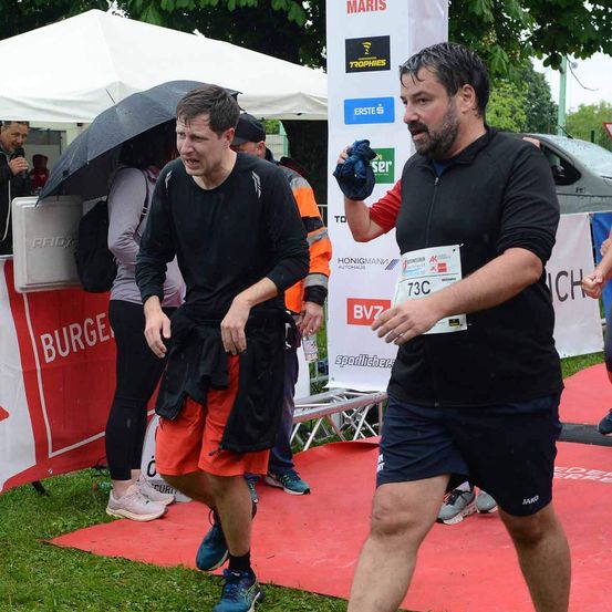 Zwei Männer laufen auf einem roten Teppich bei einem Marathon. Einer trägt ein schwarzes Shirt mit der Nummer 73C. Hinter ihnen ist ein Auto neben einem weißen Zelt geparkt.