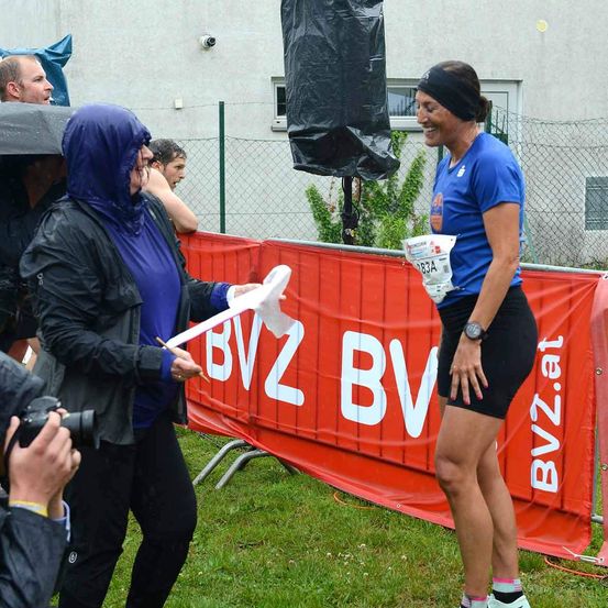 Eine Frau in einem blauen Shirt und schwarzen Shorts lächelt und hält ein Stück Papier in der Hand, während sie vor einem roten Banner mit weißen Buchstaben steht. Eine Person mit einer Kamera hält den Moment fest.
