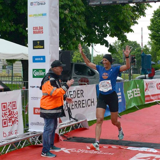 Eine Läuferin beendet einen Marathon und feiert, während ein Mann sie beglückwünscht. Die Szene ist im Freien unter einem Baum mit Bannern und Zelten im Hintergrund.