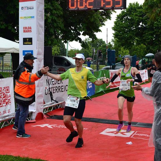 Ein Marathonläufer schüttelt die Hand eines Freiwilligen, während eine Frau vorbeiläuft. Die digitale Uhr zeigt die Zeit an. Im Hintergrund stehen Leute unter Regenschirmen.