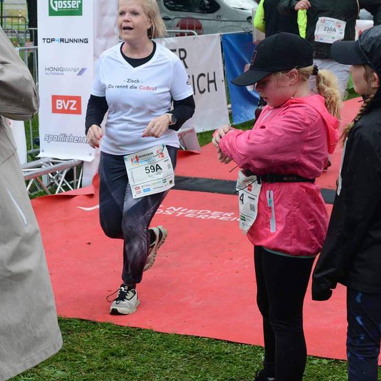 Eine Frau in einem weißen Shirt mit der Nummer 59A läuft auf einer roten Laufbahn. Sie trägt schwarze Leggings und Sneakers. Zwei junge Mädchen in schwarzen und rosa Jacken stehen in der Nähe. Im Hintergrund befindet sich ein Zaun mit Bannern.