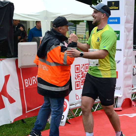 Ein Mann in einem grün-gelben Shirt schüttelt einem Mann in einem orangefarbenen Jackett auf einem roten Teppich die Hand. Dahinter steht ein weißes Zelt mit einem Banner, auf dem 'Bureau' steht.
