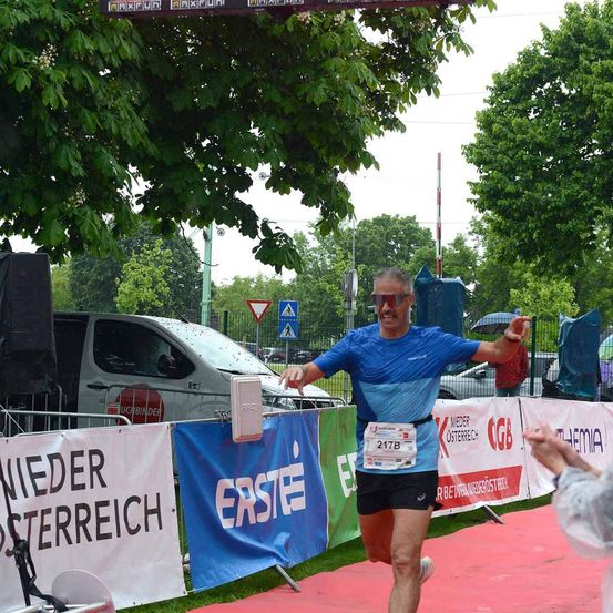 Ein Mann läuft in einem Marathon in einem blauen Shirt und schwarzen Shorts. Er befindet sich unter einem Baum und in der Nähe von Bannern. Hinter ihm steht ein Zaun mit geparkten Autos und einem Lieferwagen.