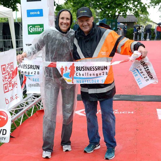 Ein Mann und eine Frau stehen auf einem roten Teppich bei einem Rennevent. Die Frau trägt eine Regenjacke und der Mann einen orangefarbenen Mantel. Sie halten ein Banner für Business Run Challenge 2B. Im Hintergrund sind Menschen und Regenschirme zu sehen.