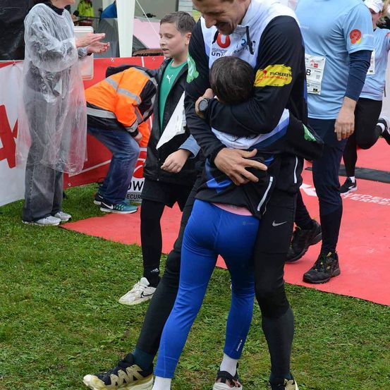 Ein Mann umarmt ein Kind in blauen Leggings bei einem Rennen, in der Nähe einer Frau in einer Regenjacke und eines Jungen im grünen Shirt. Ein weiterer Läufer in einem blauen Shirt läuft auf der Bahn.
