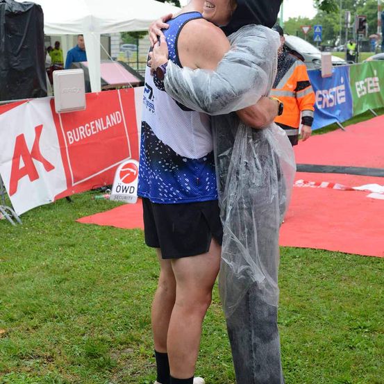 Zwei Menschen umarmen sich in einem Park. Einer trägt ein blaues und weißes Sporttrikot. Ein roter Banner mit weißen Buchstaben steht für 'Burgenland' und 'AK'. Ein orangefarbener Weste ist im Hintergrund zu sehen.