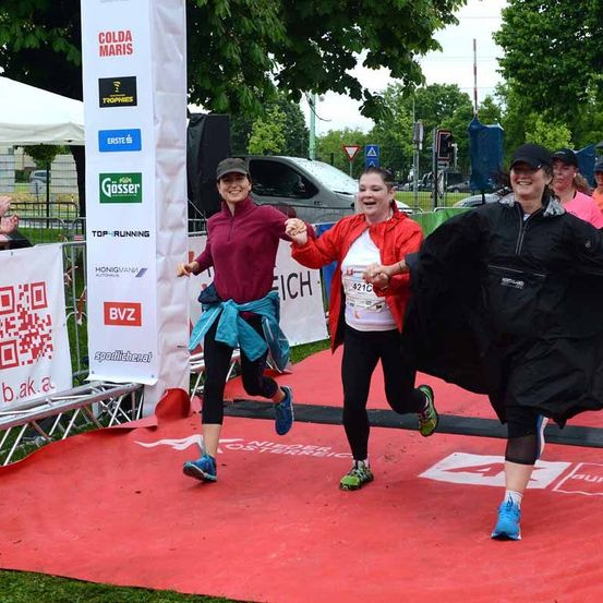 Drei Frauen laufen auf einem roten Teppich und tragen verschiedene Sportbekleidung. Sie befinden sich in einer Parkumgebung mit Bannern und Zelten. Die Frau in der Mitte trägt eine rote Jacke.