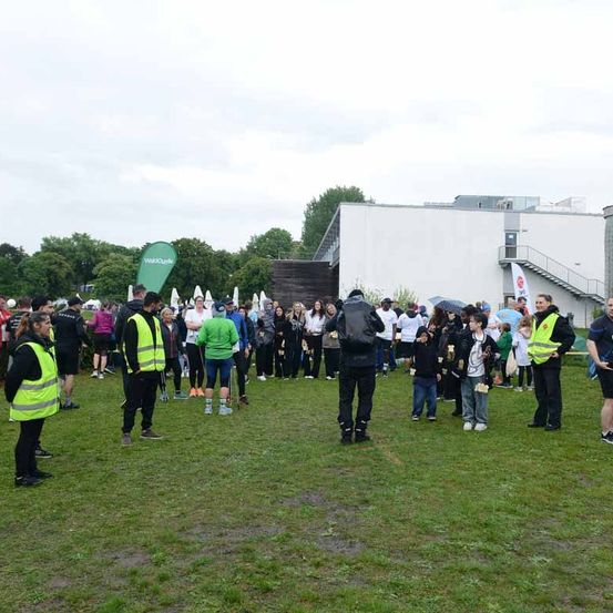 Eine Gruppe von Menschen in Warnwesten steht auf einem Rasen. Einige halten Regenschirme. Dahinter steht ein Gebäude mit Treppen und Bannern. Bäume und ein bewölkter Himmel sind im Hintergrund.