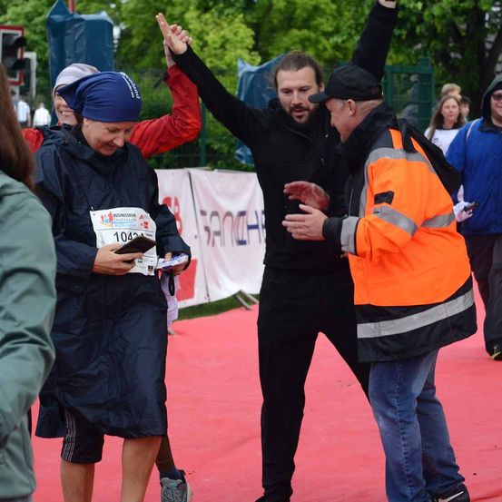 Eine Frau mit einem blauen Kopftuch und der Nummer 1042 auf ihrem Shirt unterhält sich mit zwei Männern auf einem roten Teppich. Ein Mann trägt eine Warnweste und ein anderer Mann hebt seinen Arm.
