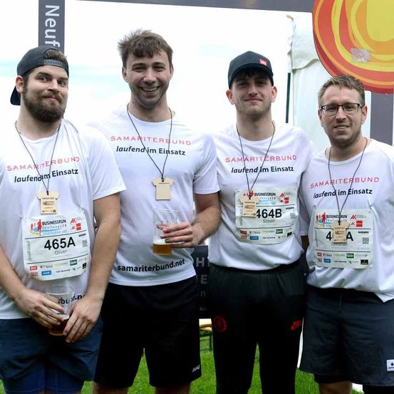 Vier Männer, die weiße T-Shirts mit Samaritan Association tragen, stehen nebeneinander und posieren möglicherweise für ein Foto. Sie halten alle Getränke in den Händen.